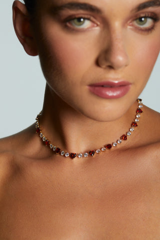 MODEL WEARING LE BISOU RED HEART COLLAR NECKLACE. HER FACE IS OUT OF FOCUS AND IT IS A CLOSE UP OF HER NECKLINE.