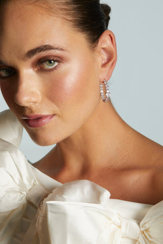 Close-up of a woman wearing LE BISOU HEART C HOOP EARRINGS with a white bow dress against a light blurred background