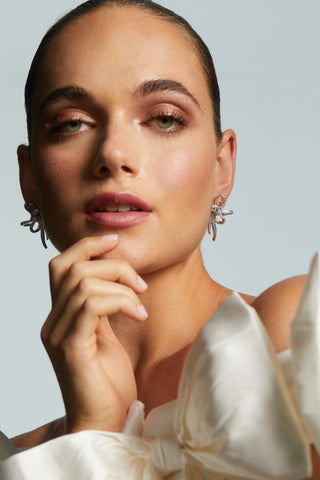 Woman wearing LE BISOU PAVE BOW STUD EARRINGS with a neutral background and white puffy bow dress