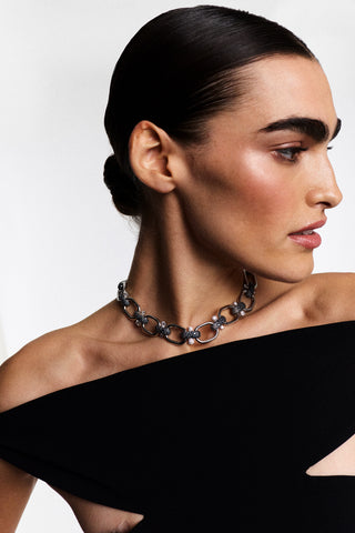 A MODEL TURNED TO THE SIDE WEARING A BLACK DRESS, HAIR IN A SLICK-BACK BUN, AND THE TURN UP THE VOLUME LINK ALL AROUND NECKLACE