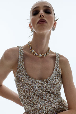 A MODEL STANDING WITH HER EYES CLOSED AS SHE WEARS A GOLD SPARKLY DRESS AND THE TURN UP THE VOLUME LINK ALL AROUND NECKLACE