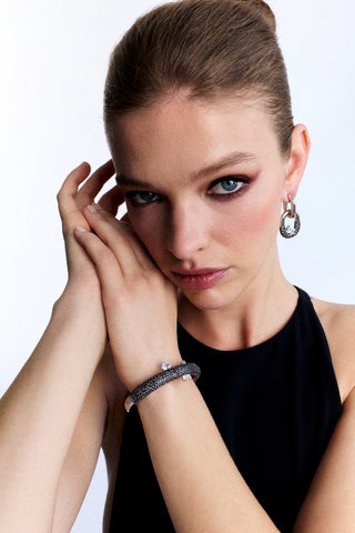 MODEL LOOKING AT THE CAMERA WITH HER HANDS UP RESTING AGAINST HER FACE, AS SHE WEARS A BLACK TOP AND THE TURN UP THE VOLUME SIDE HINGE BRACELET