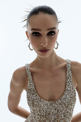 A MODEL LOOKING AT THE CAMERA AS SHE WEARS SULTRY MAKEUP, A GOLD SPARKLY DRESS, AND THE TURN UP THE VOLUME LARGE HOOP EARRINGS