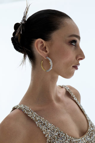 SIDE PROFILE OF A MODEL WEARING A GOLD SPARKLY DRESS, THE TURN UP THE VOLUME LARGE HOOP EARRINGS, AND HER HAIR IN A SPIKY SLICK-BACK BUN