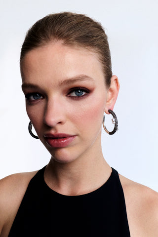 A MODEL LOOKING AT THE CAMERA WITH SULTRY MAKEUP WEARING A BLACK HIGH-NECK DRESS AND THE TURN UP THE VOLUME LARGE HOOP EARRINGS