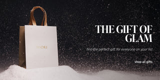 White gift bag with gold handle on snow, 'NADRI' brand visible, text 'The Gift of Glam' and 'shop all gifts' on dark snowy background.