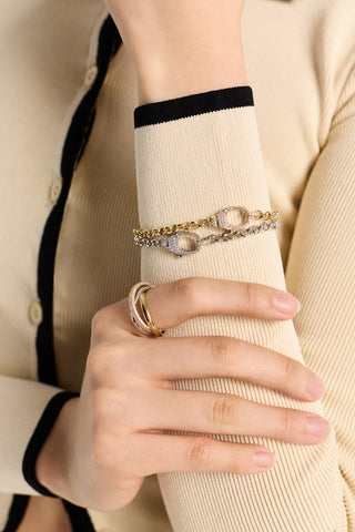 CLOSE UP OF A MODEL RAISING HER WRIST AND HOLDING HER ARM, WEARING AN IVORY SHIRT AND GEMINI PAVE CLASP LINE BRACELETS IN BOTH GOLD AND RHODIUM PLATING OVER HER SLEEVE