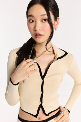 MODEL WEARING AN IVORY TOP WITH BLACK TRIM AND THE GEMINI PAVÉ CLASP NECKLACE, LOOKING OFF TO THE SIDE AND HOLDING THE NECKLACE WITH HER HAIR FRAMING HER FACE
