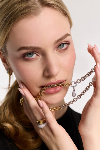 CLOSE UP OF A MODEL LOOKING AT THE CAMERA, HOLDING THE GEMINI PAVE DROP CHAIN NECKLACE IN BETWEEN HER TWO HANDS AND PART OF THE CHAIN IN HER MOUTH. 