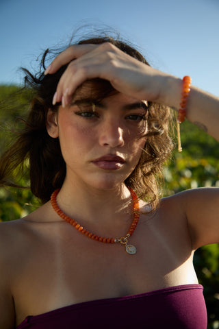 MODEL OUTSIDE WITH HER HAIR BLOWING AND HAND LIFTED TO HER FOREHEAD, WEARING THE BEADS OF PARADISE SUN PENDANT NECKLACE AND MATCHING BOLO BRACELET 