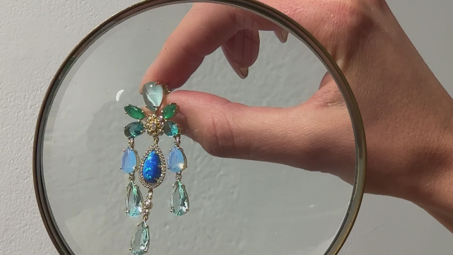 video shows a model carefully examining Nadri's Blue Lagoon chandelier earrings as well as seashells under a magnifying glass. 