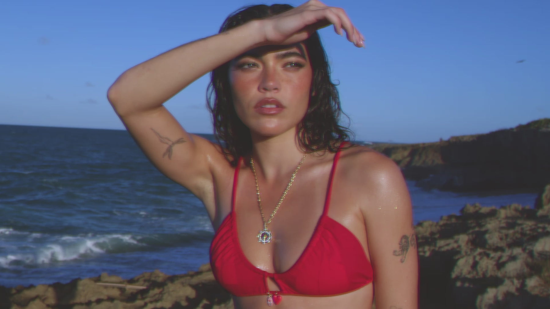 Video of model in a red bikini wearing the GlobeTrotter Compass pendant while on a rocky beach.