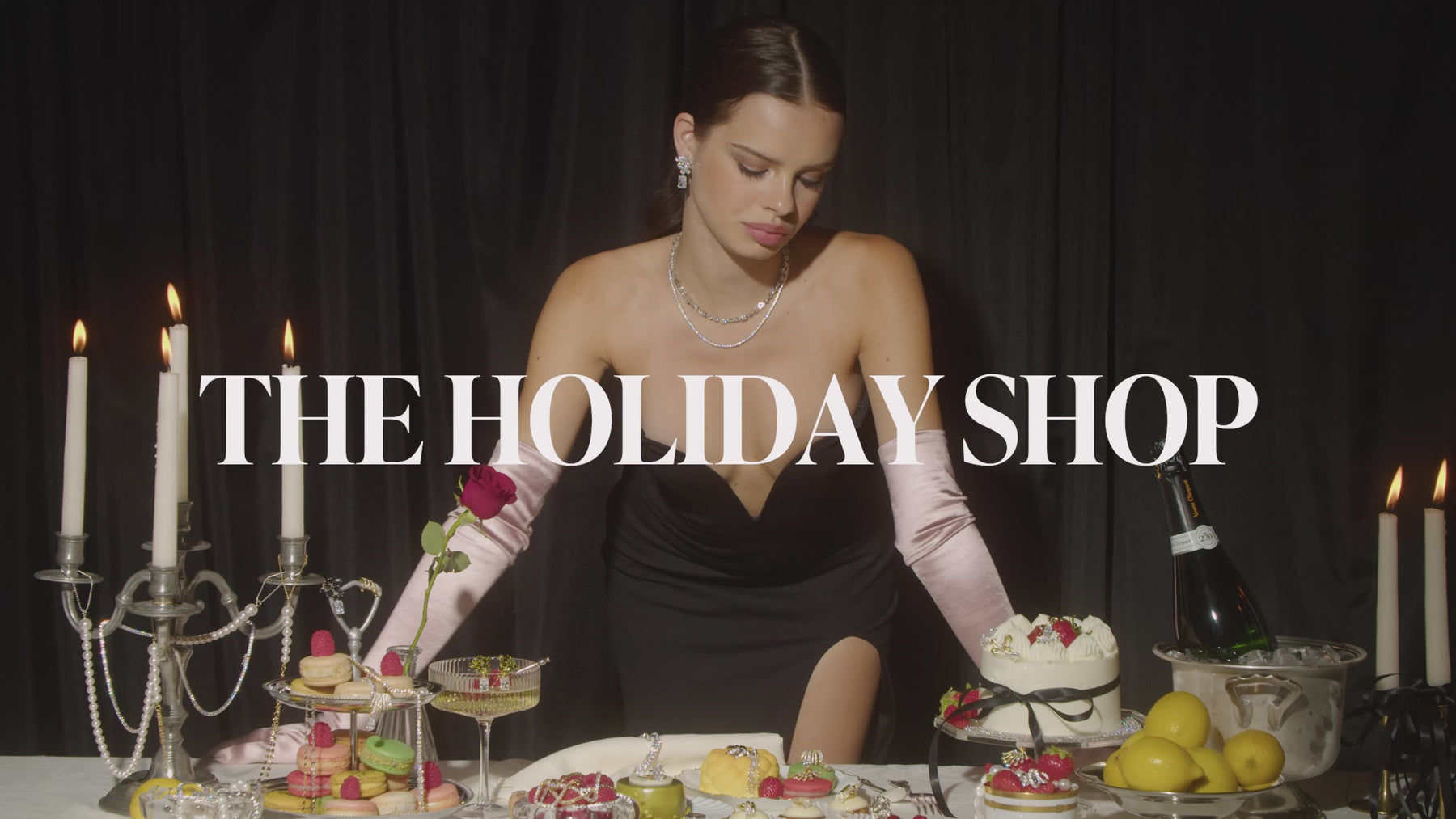 Video of a model in a black evening gown with pink satin gloves and Nadri jewelry standing in front of a table that is covered with desserts and more Nadri jewelry.  The model is playfully showing off the jewelry, laughing, and looking into the camera. The vibe is moody but festive and celebratory. Text says "The Holiday Shop".